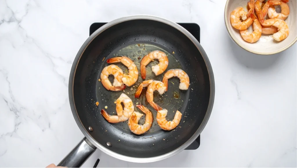 Shrimp sautéing in sesame oil in a stainless steel skillet on a white marble kitchen countertop, a key step in making Korean glass noodles