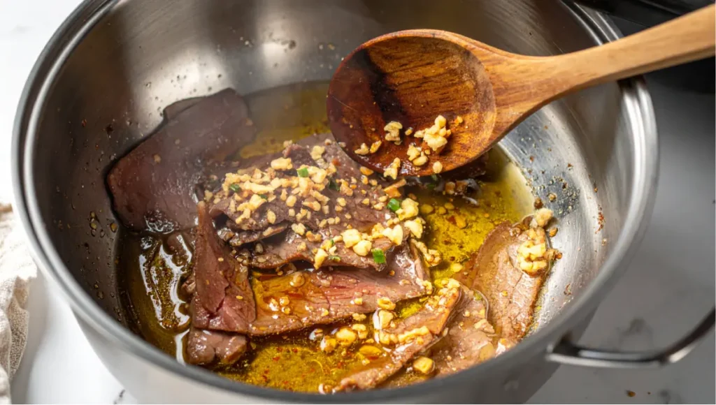 Close-up of beef brisket slices and minced garlic sautéing in sesame oil in a stainless steel pot on a white marble countertop during Korean beef soup preparation