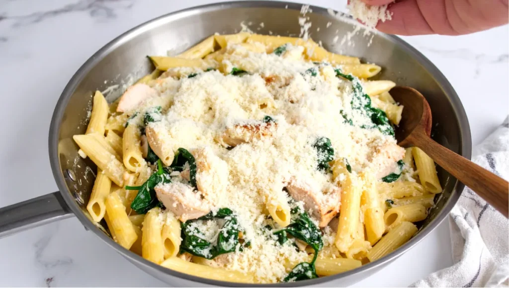 Creamy chicken spinach pasta in a stainless steel skillet with Parmesan being scattered on top, resting on a white marble kitchen countertop